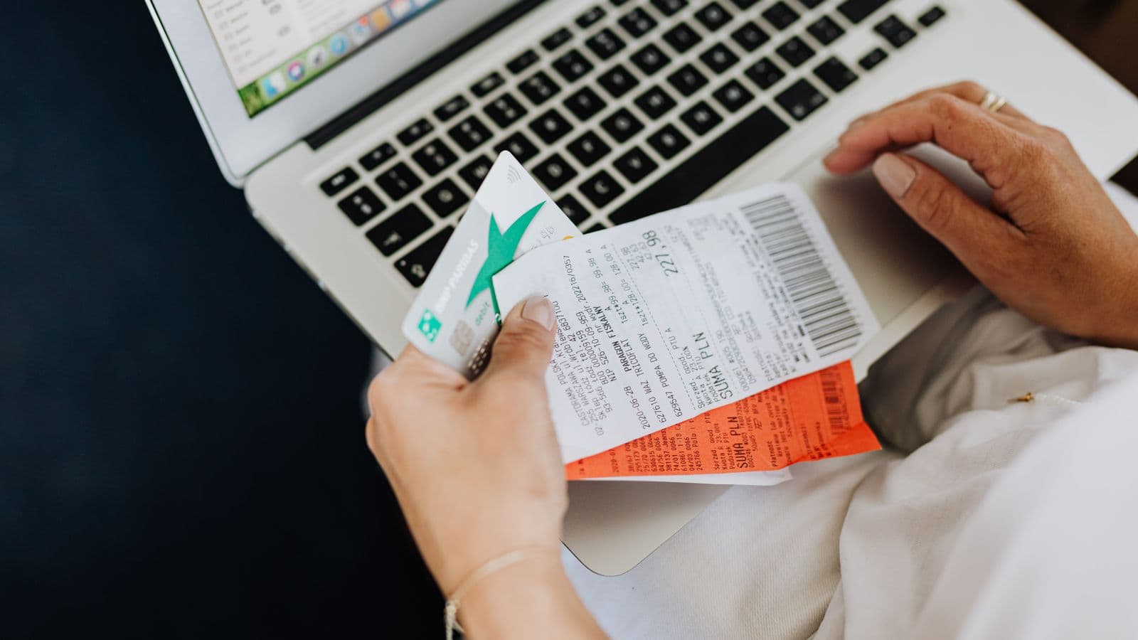 Close-up of hands holding receipts and a bank card in front of a laptop while reconciling credit card statements